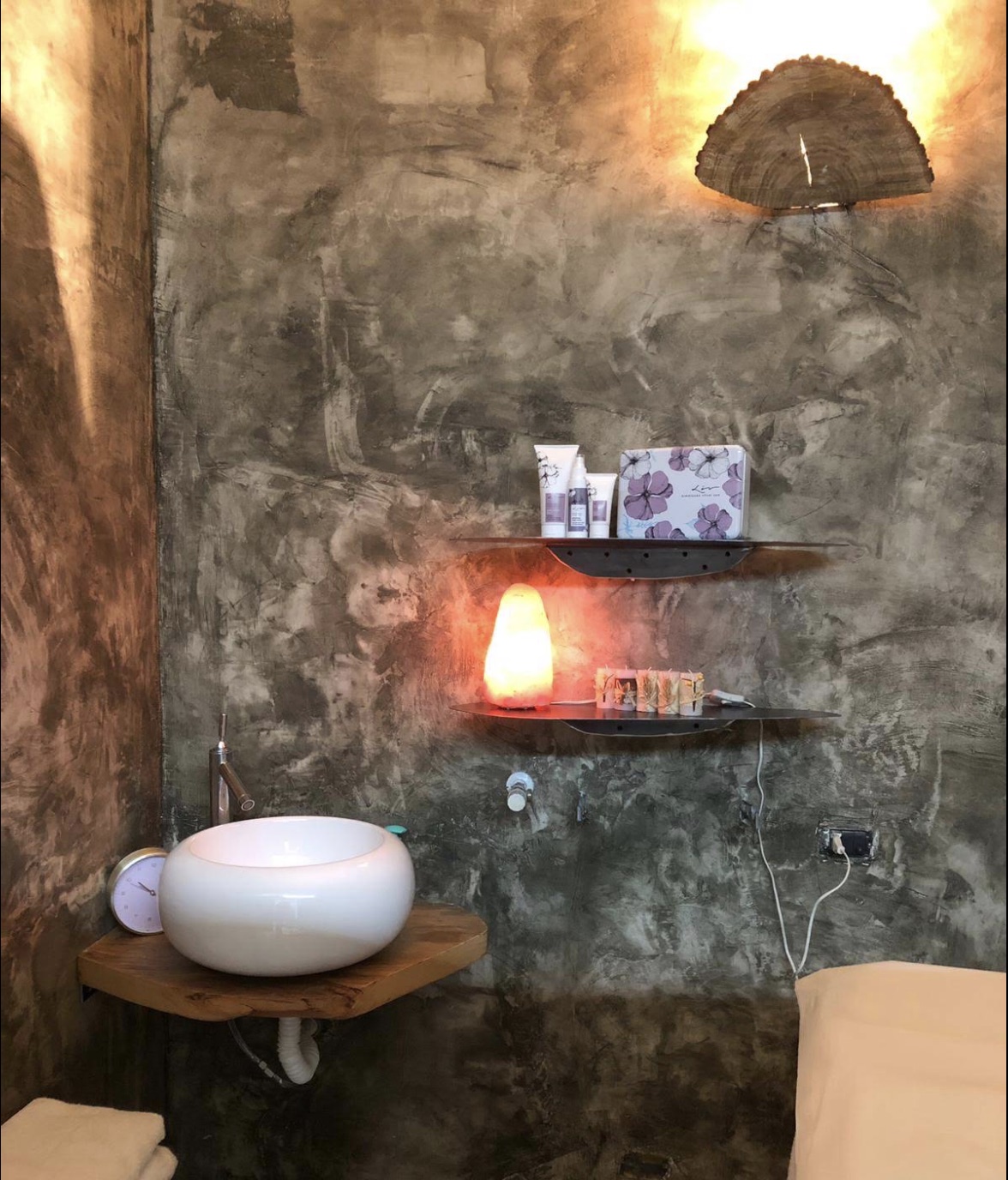 Una cabina Rosemery: lavabo in ceramica su mensola in legno, lampada di sale e luce calda su parete materica